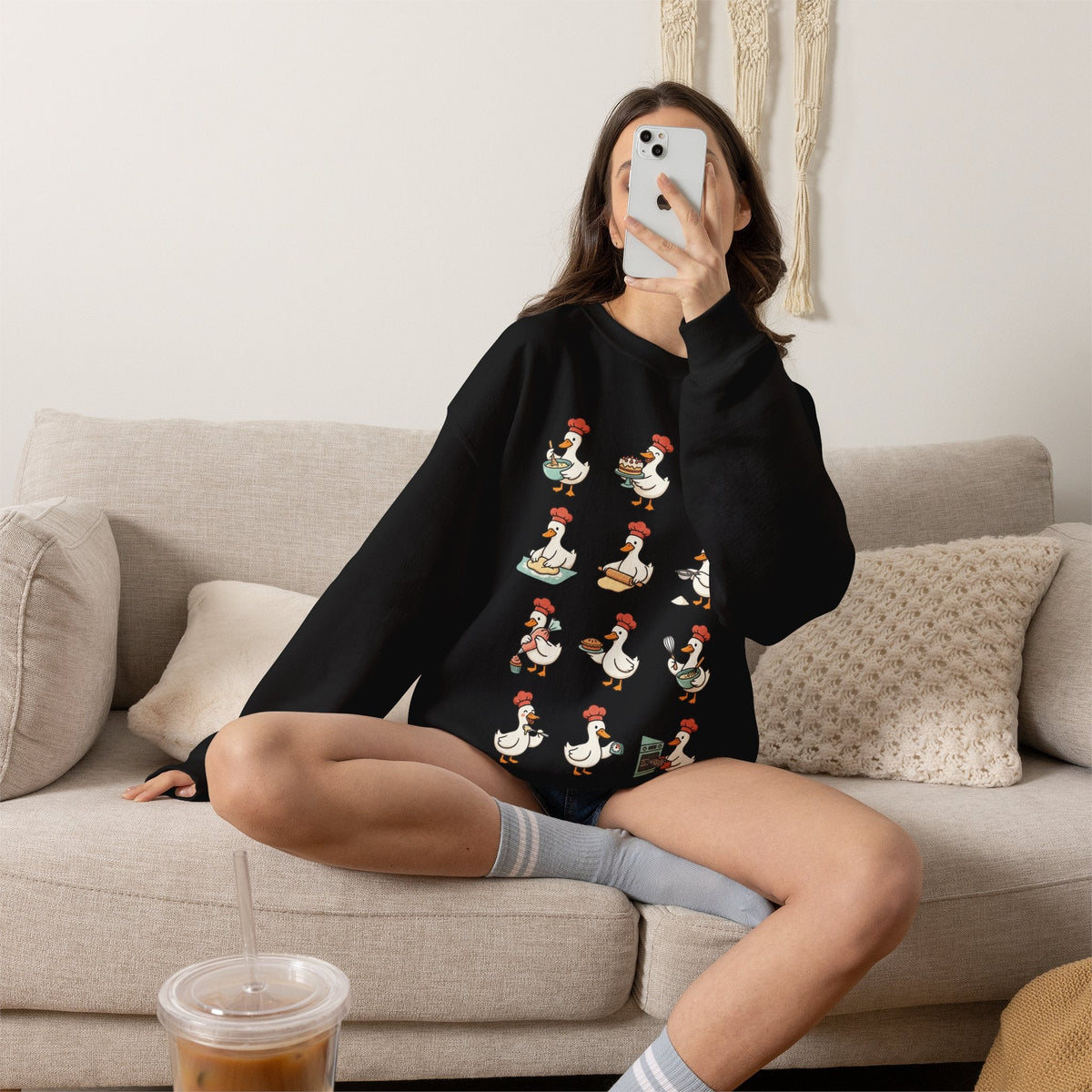 Wicked Thread Society black crewneck sweatshirt featuring a whimsical chef goose print, worn by a woman on a sofa.