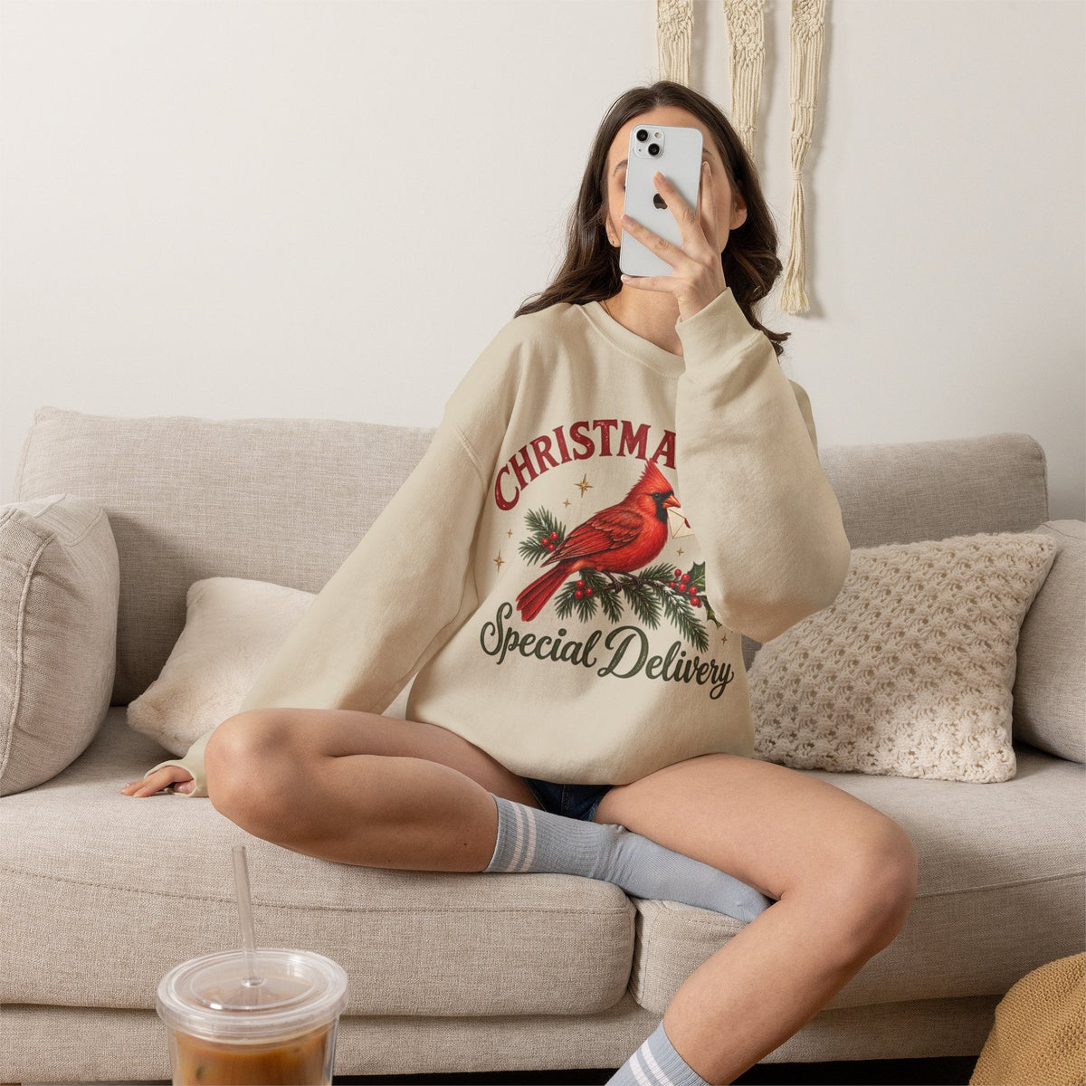 Wicked Thread Society beige crewneck sweatshirt with a cardinal Christmas post graphic, worn by a woman on a sofa.