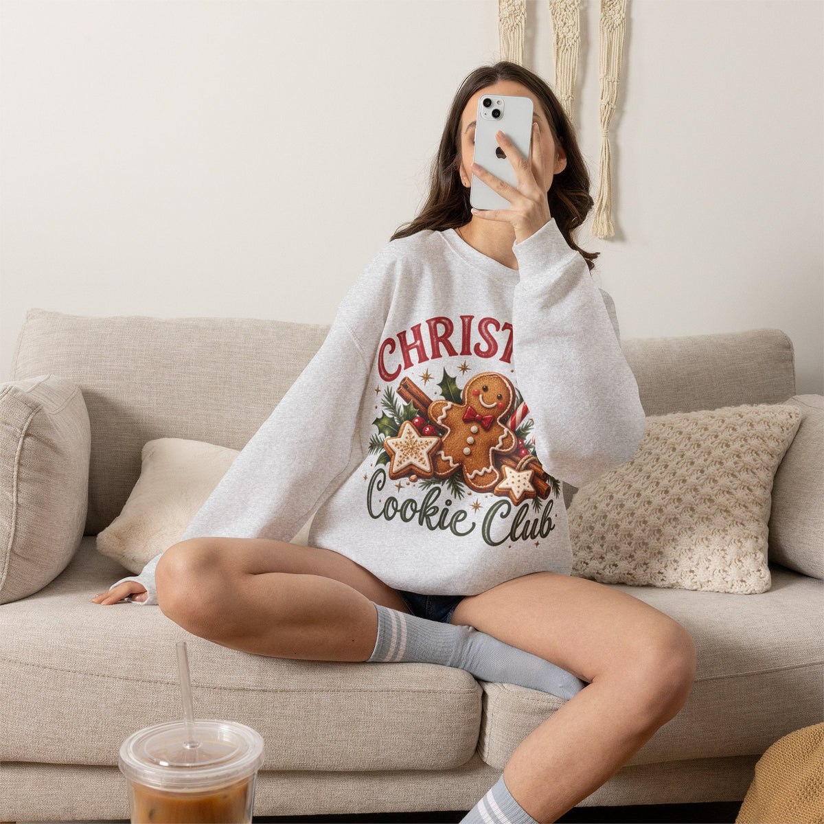 Wicked Thread Society grey crewneck sweatshirt featuring a gingerbread man graphic, worn by a woman on a beige sofa.