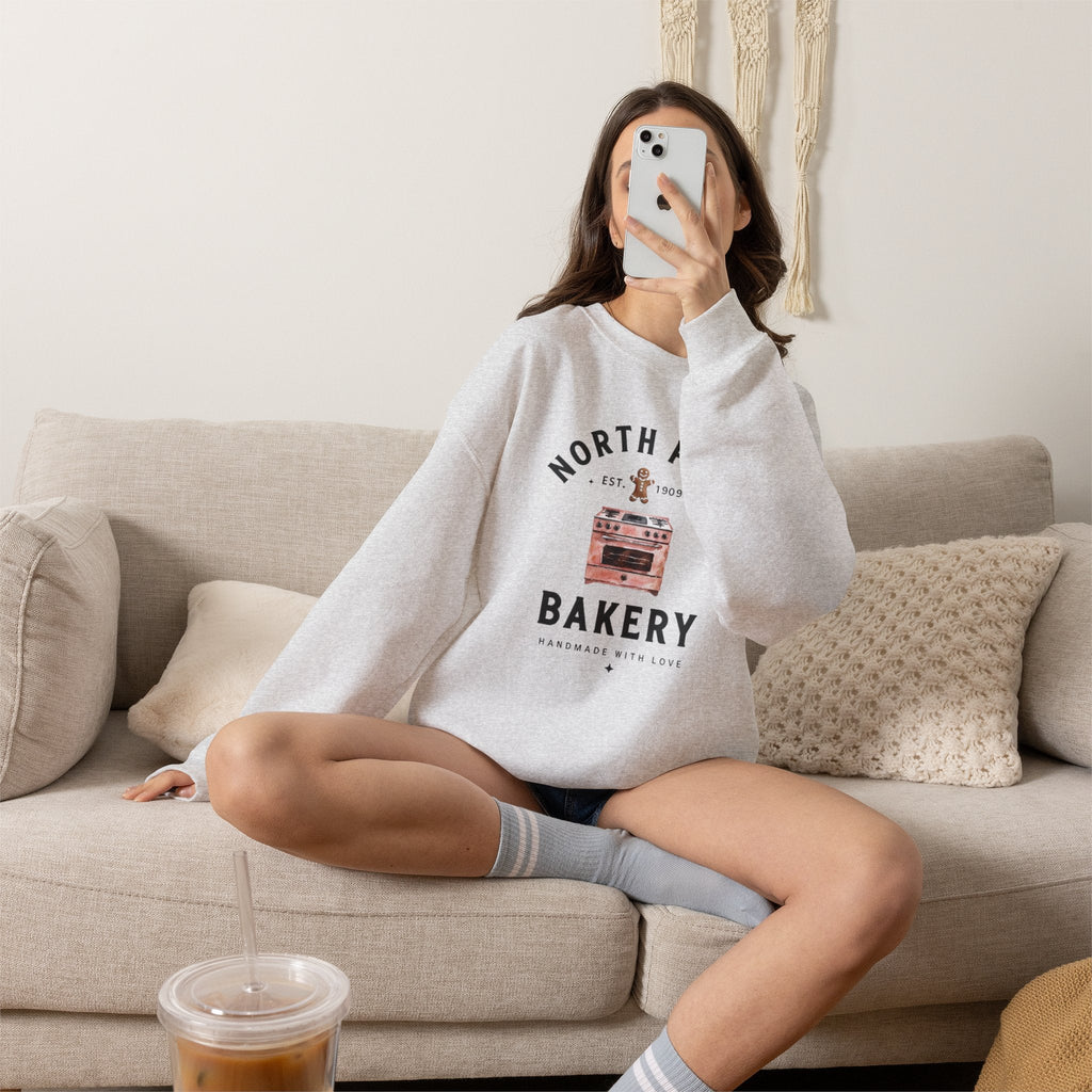 Wicked Thread Society grey crewneck sweatshirt with North Pole Bakery oven and gingerbread man graphic on a couch