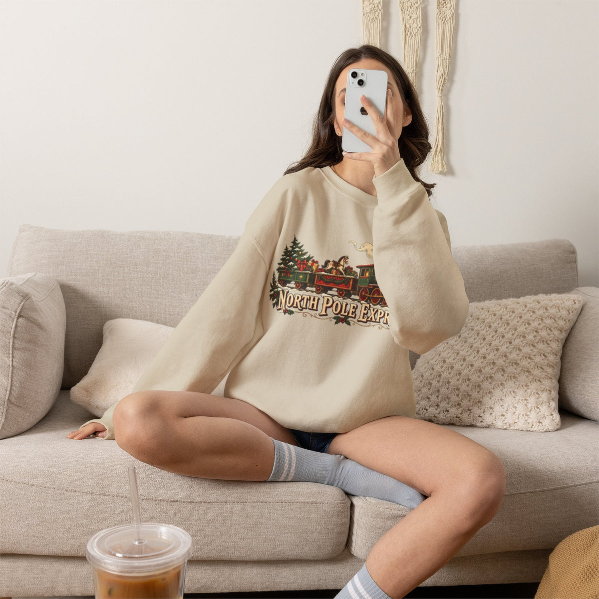 Wicked Thread Society beige crewneck sweatshirt with vintage North Pole Express train graphic worn by a woman on a sofa