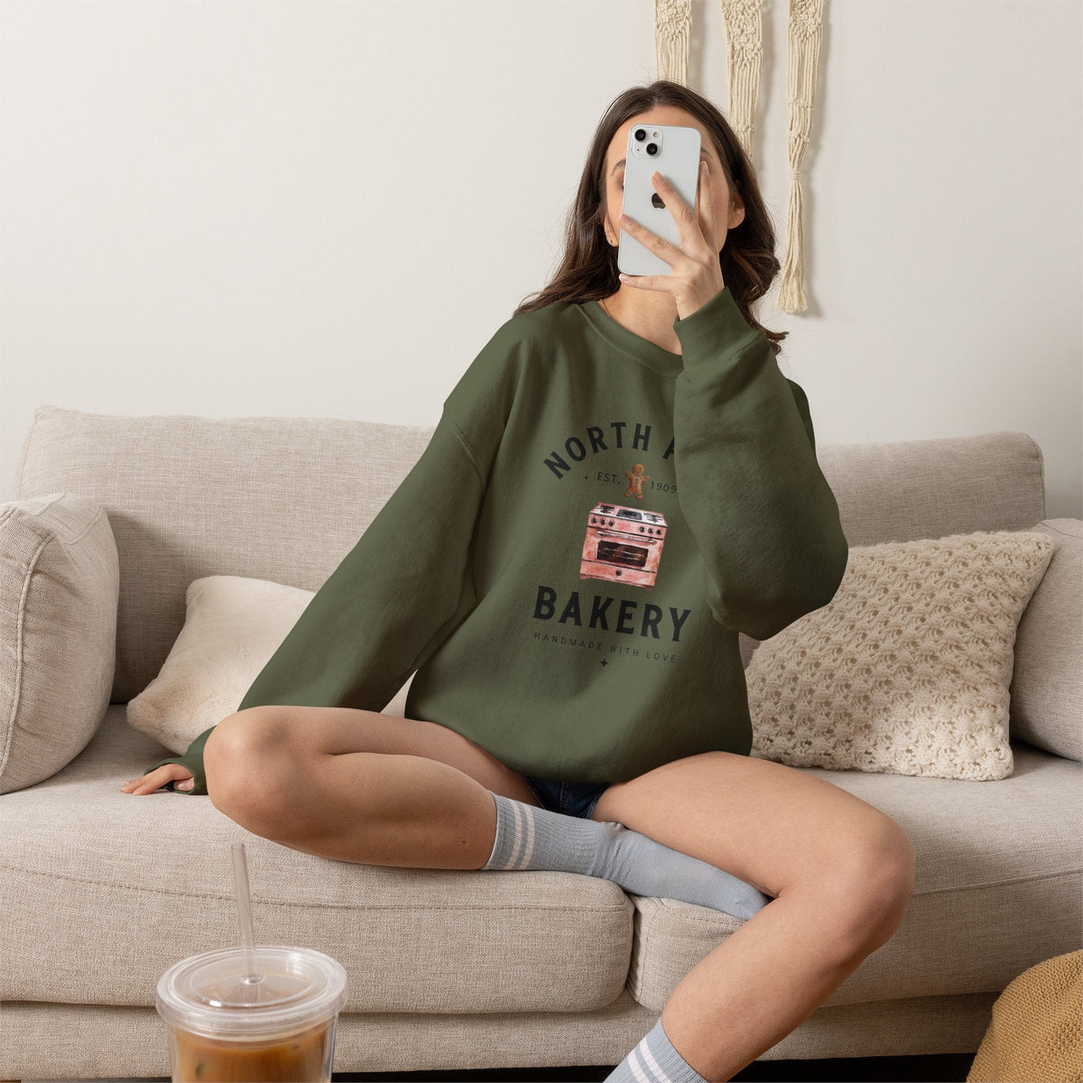 Wicked Thread Society olive green crewneck sweatshirt with North Pole Bakery oven graphic worn by a woman on a sofa.