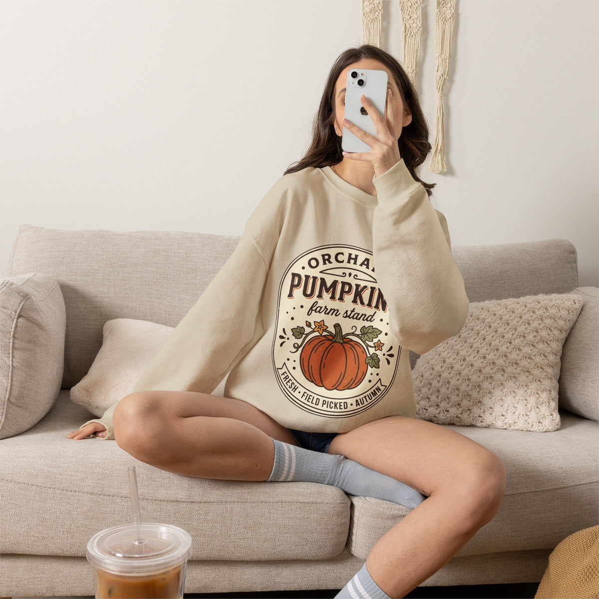 Wicked Thread Society beige crewneck sweatshirt with a vintage pumpkin farmstand graphic worn by a woman on a sofa.