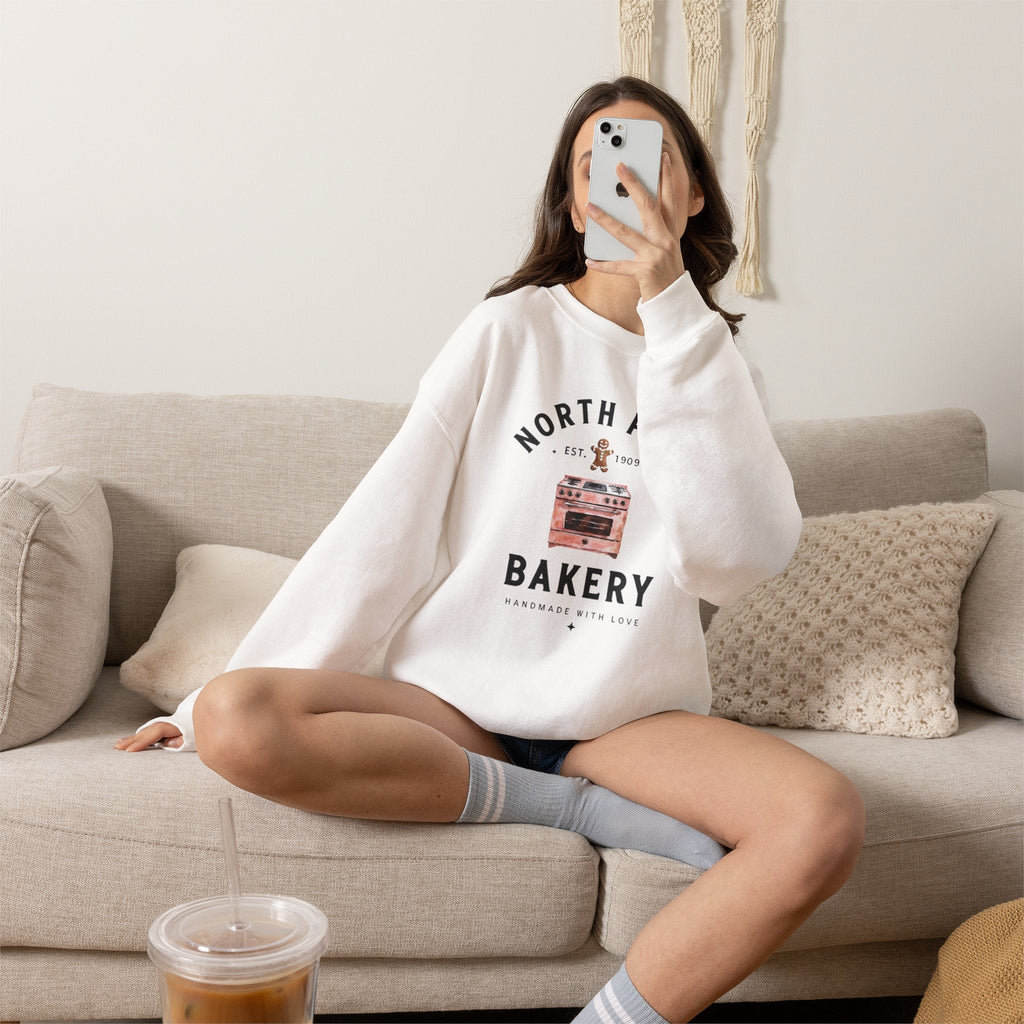 Wicked Thread Society white crewneck sweatshirt with North Pole Bakery oven and gingerbread man graphic on a sofa